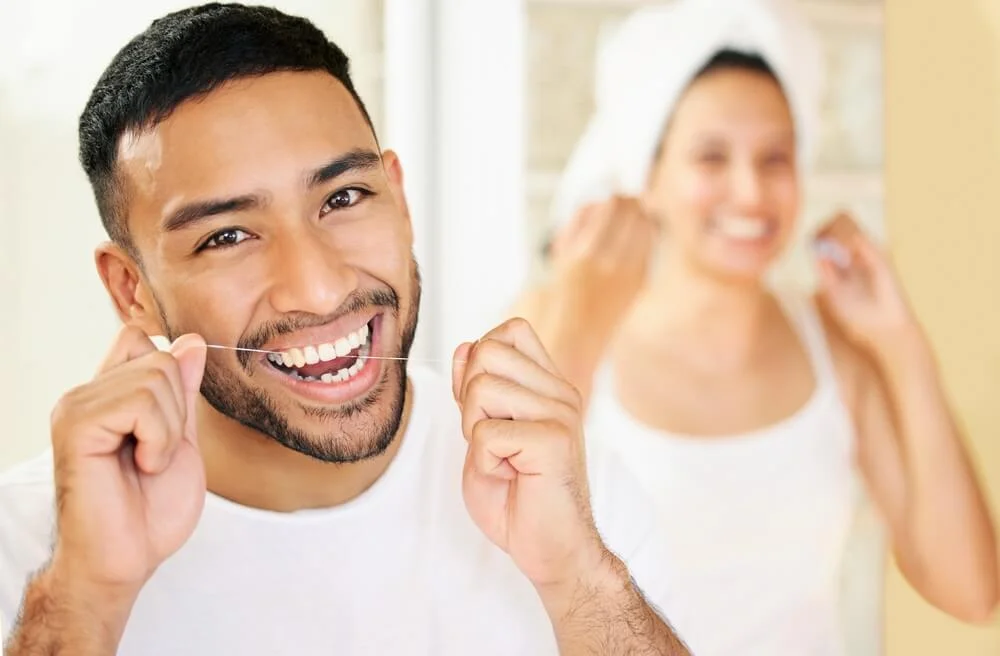 Man flossing teeth with a woman blurred in the background.